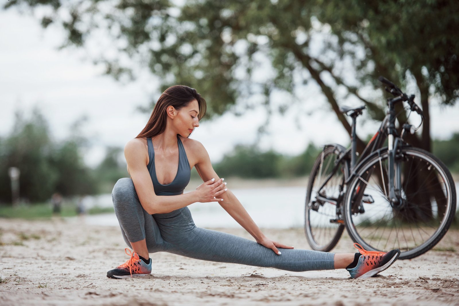 Concentrated look. Female cyclist with good body shape doing yoga exercises and stretching near her bike on beach at daytime.